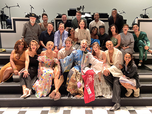 Shechter sits smiling in the Robe in the middle of a group shot of about 23 people on steps on a stage in front of a visible orchestra area.