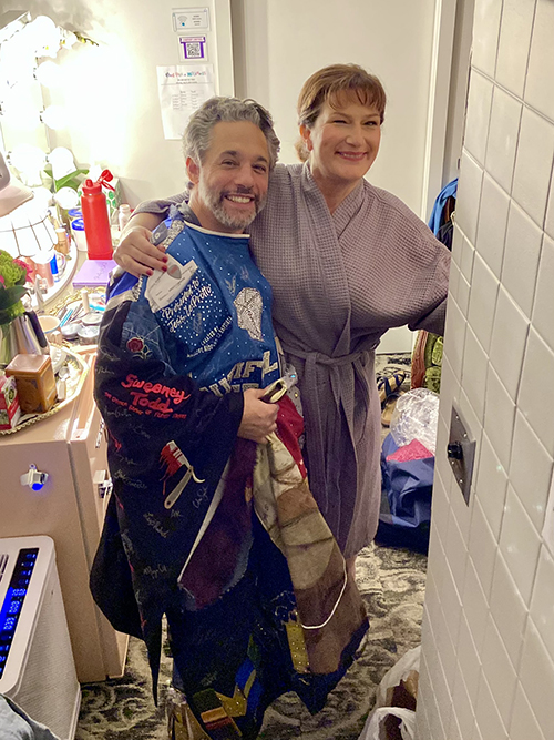 Schecter wears the Robe backstage. Ana Gasteyer, in a dressing robe, puts her arm around him. Both are smiling.