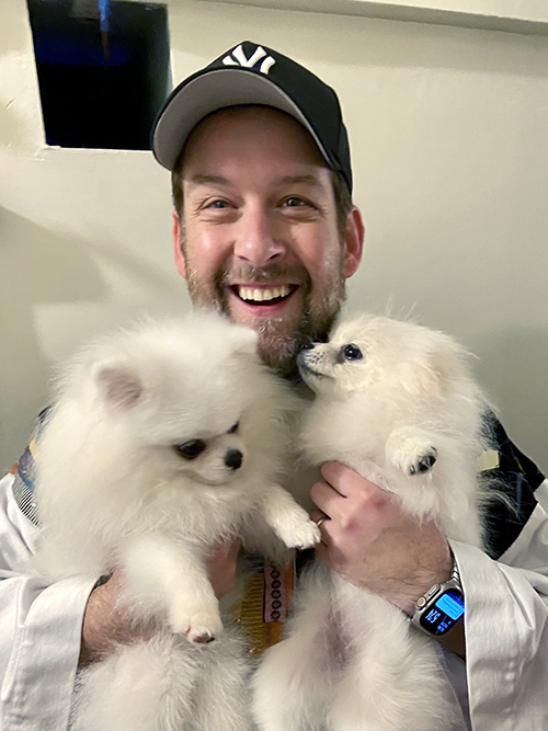 Kober smiles in the Robe while holding up two small fluffy white dogs.