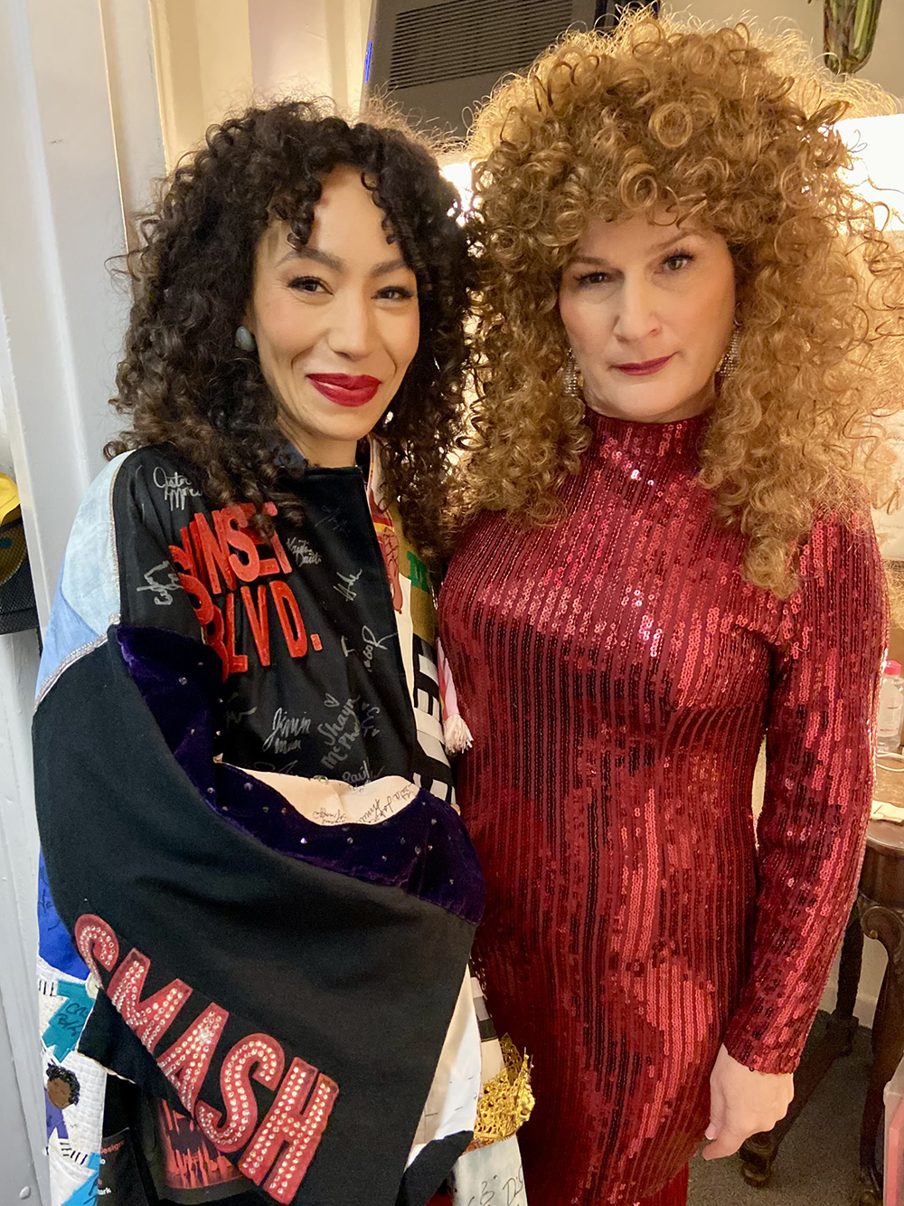 Afra Hines stands in the Robe next to Ana Gasteyer, who is wearing a red sequined gown and redhead wig with tight curls.