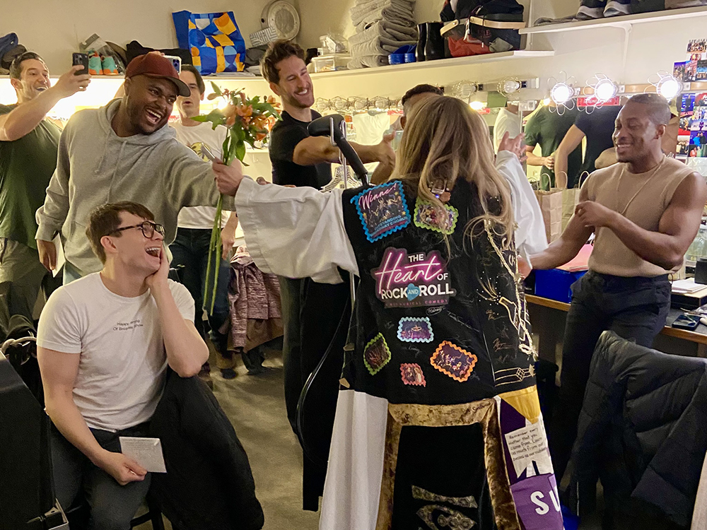 Webber's back is visible as she stretches out her arms in the Robe, holding flowers in one hand, in a dressing room, towards about seven castmates who look thrilled.